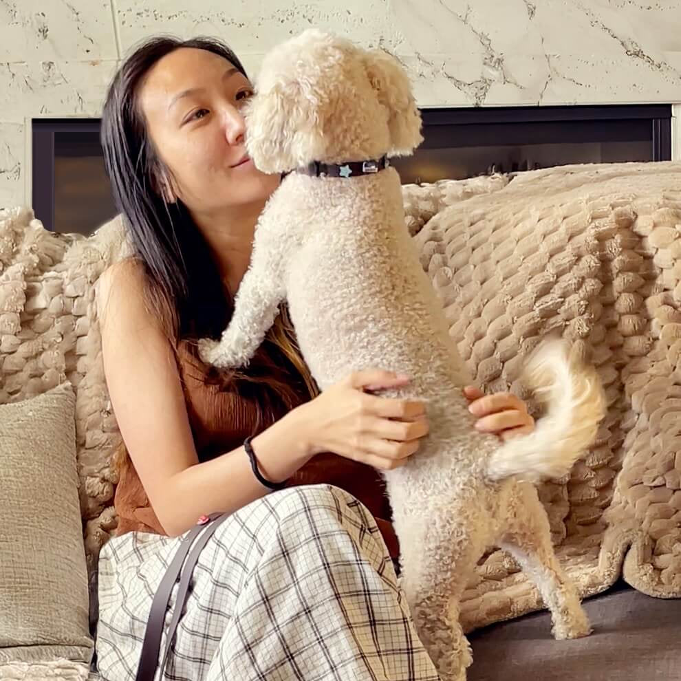 Woman holding a small white dog wearing a Pooches & Cream custom collar with dog collar charms on a couch in a cozy living room.
