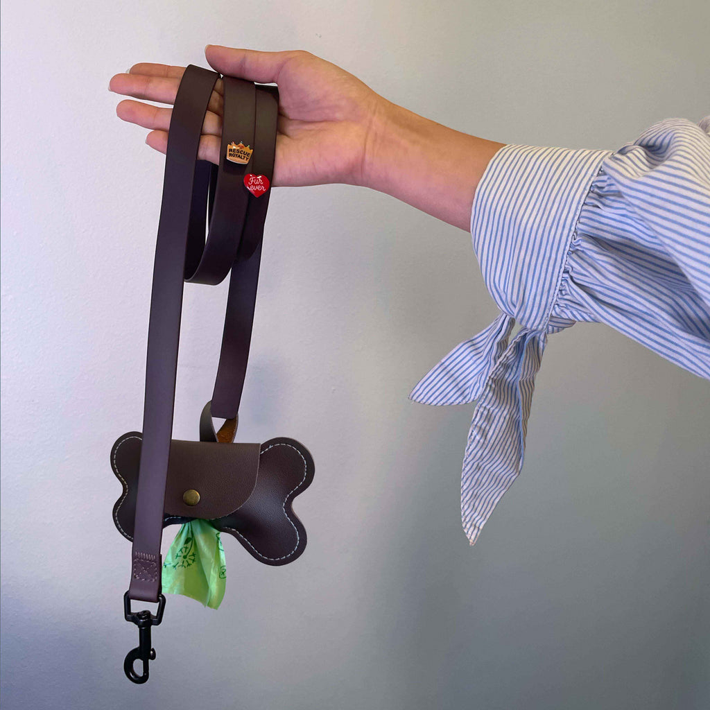 Hand holding a vegan leather dog leash with a bone shaped matching bag against a plain background.